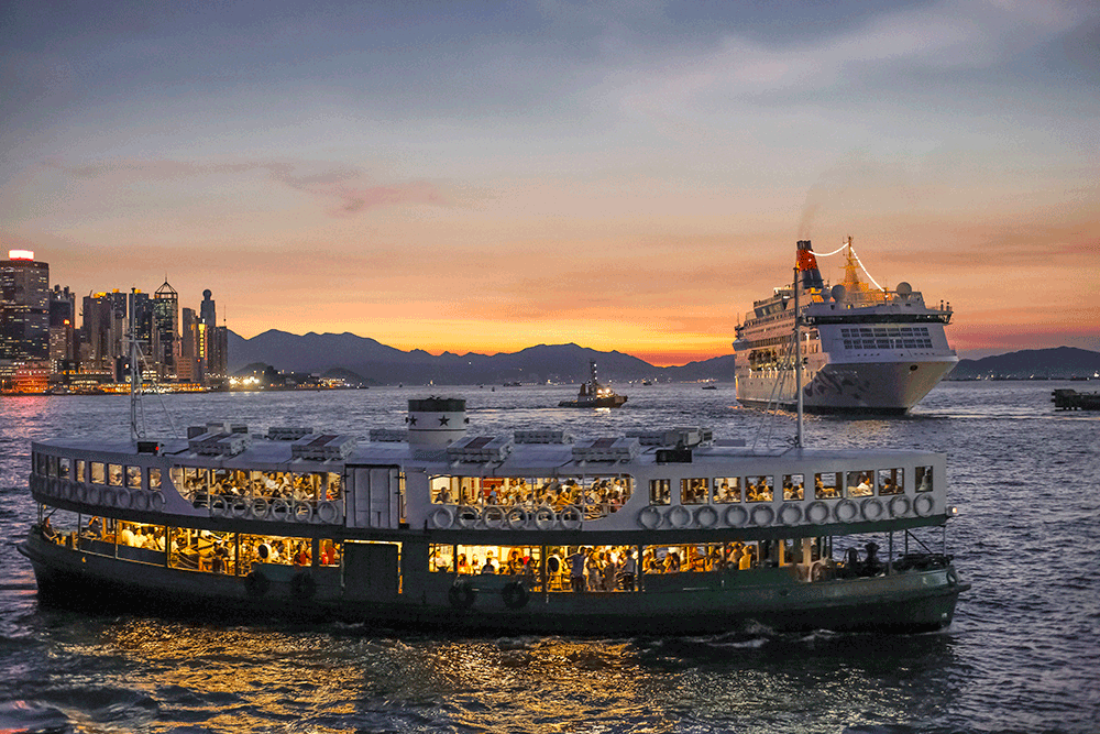 A double-decker ferry with illuminated interiors is sailing across Victoria Harbour at sunset, with a larger cruise ship and a city skyline in the background.